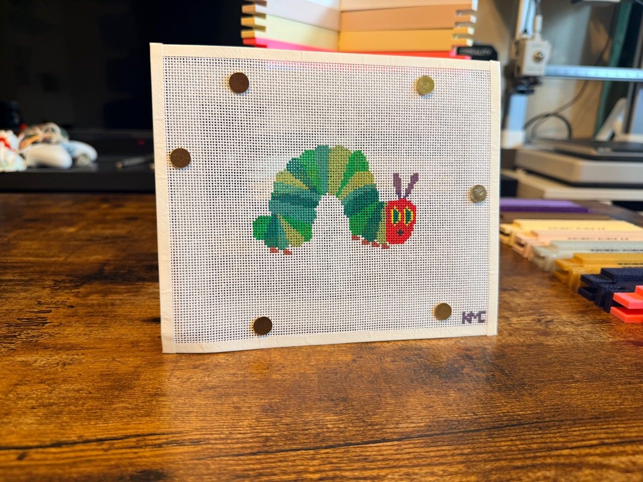A stitched needlepoint canvas mounted on a white Denton Curio Co stretcher bar frame